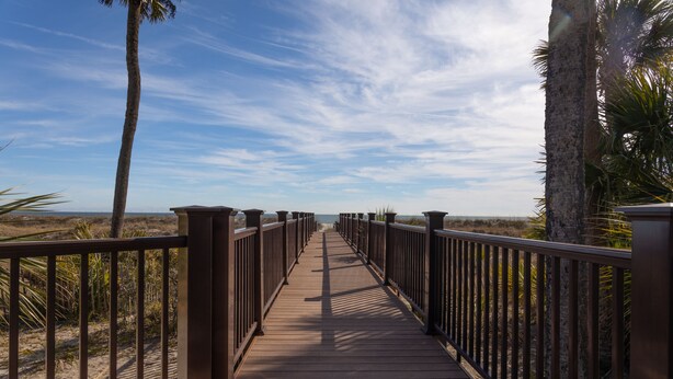 Un passeig de fusta amb baranes metàl·liques que condueix a una platja amb palmeres i un cel serè.