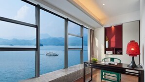 A room with a large window overlooking a lake and mountains, a desk with a chair, and a red lamp. - Qiandaohu Kaiyuan Life Hotel (Chun'an)
