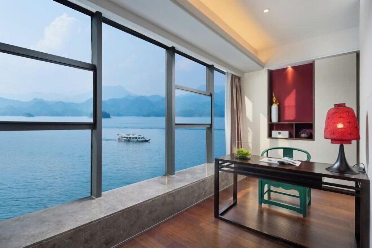 Ein Zimmer mit großem Fenster mit Blick auf ein Gewässer und Berge, ein Schreibtisch mit Stuhl und eine rote Lampe.