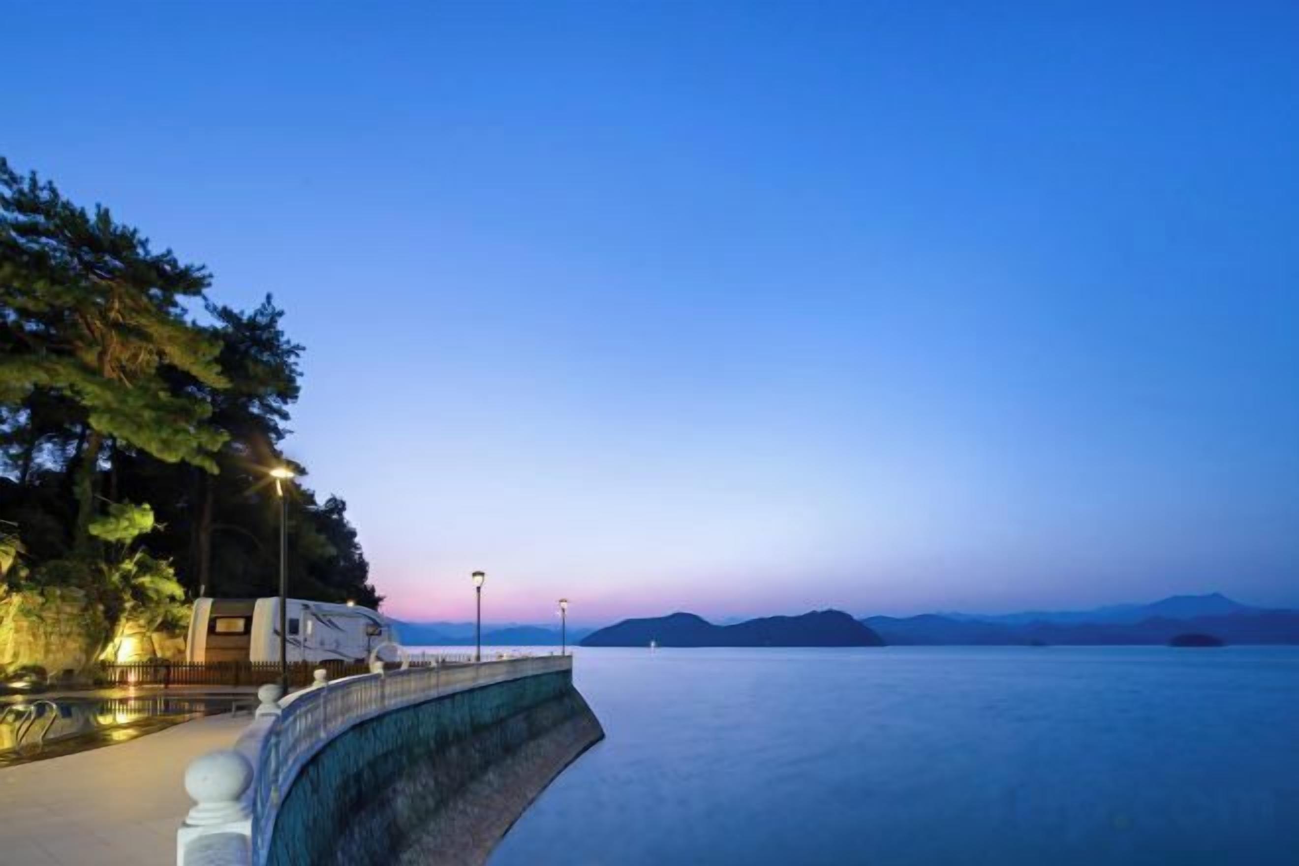 A waterfront property with a curved stone wall, a parked RV, and a row of street lamps.