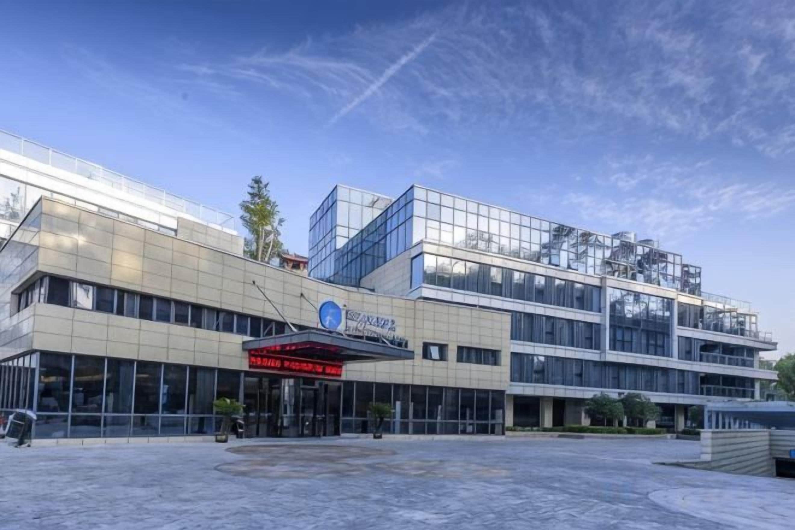 A modern building with a glass facade, a prominent entrance with a red sign, and a clear sky.