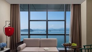 A room with a sofa, a small table with a bowl of fruit, and a view of the sea through large windows. - Qiandaohu Kaiyuan Life Hotel (Chun'an)