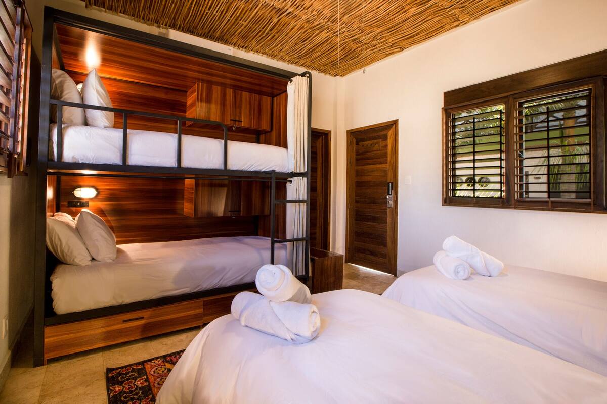 A room with bunk beds, a wooden ceiling, a window with blinds, and a door.