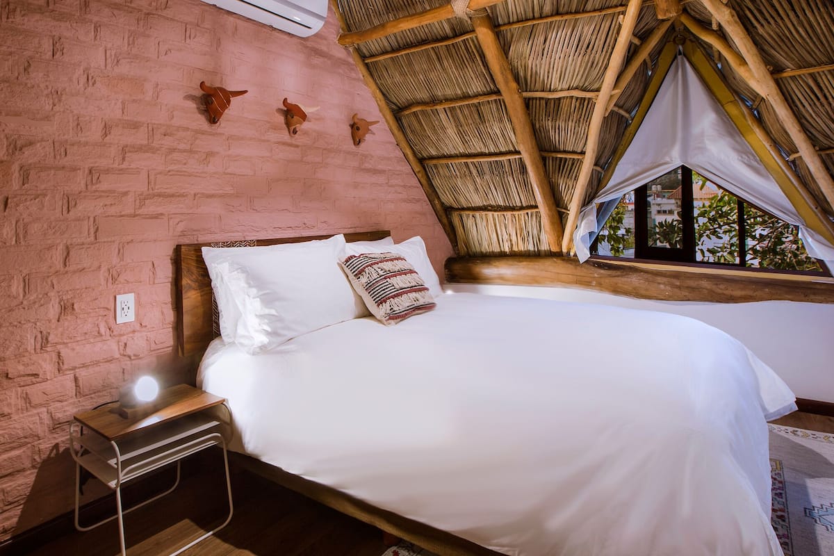 A bedroom with a brick wall, a bed with white bedding, a nightstand, and a window with a thatched roof.