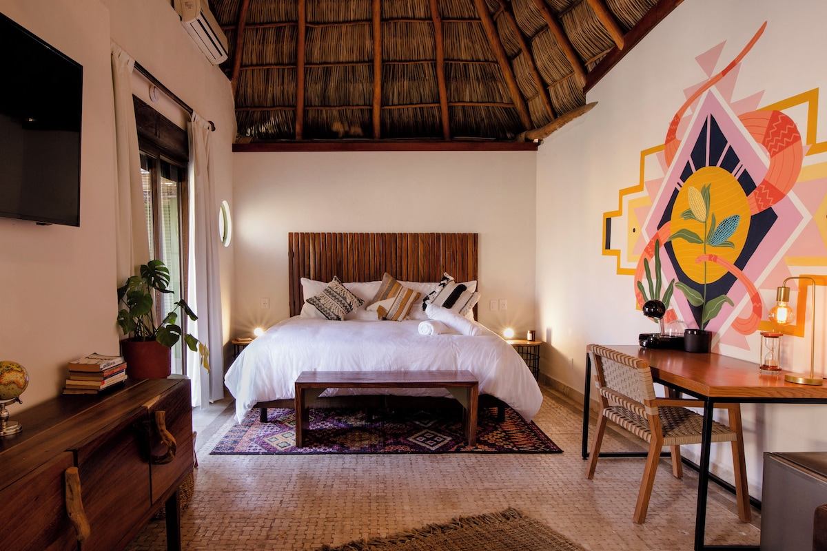 A bedroom with a bed, bedside tables, a desk, a chair, a potted plant, and a wall with a geometric design.