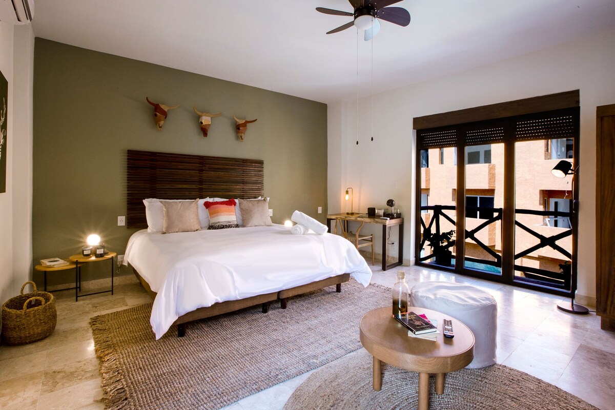 A well-lit bedroom with a large bed, bedside tables, a ceiling fan, and a view through a glass door to an outdoor area.