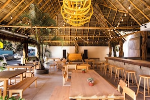 An indoor dining area with wooden tables and chairs, a thatched roof, and string lights.