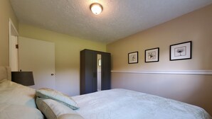 Un dormitorio con cama, almohadas, armario, mesita de noche y cuadros en la pared.