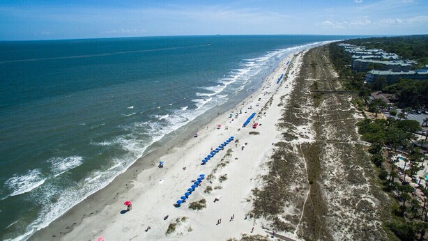 An aerial view of a beach with rows of umbrellas, a long stretch of beach, and buildings in the background.
