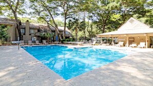 A swimming pool with lounge chairs and a covered area, surrounded by trees and a building with a sloped roof.