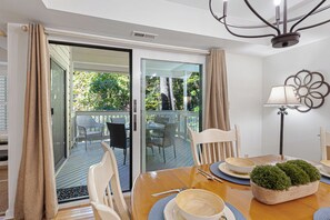 A dining area with a table set for two, a chair, a lamp, and a view through a glass door to an outdoor deck.