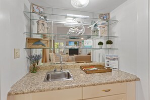A kitchen with a granite countertop, stainless steel sink, and a glass shelf unit above the sink.