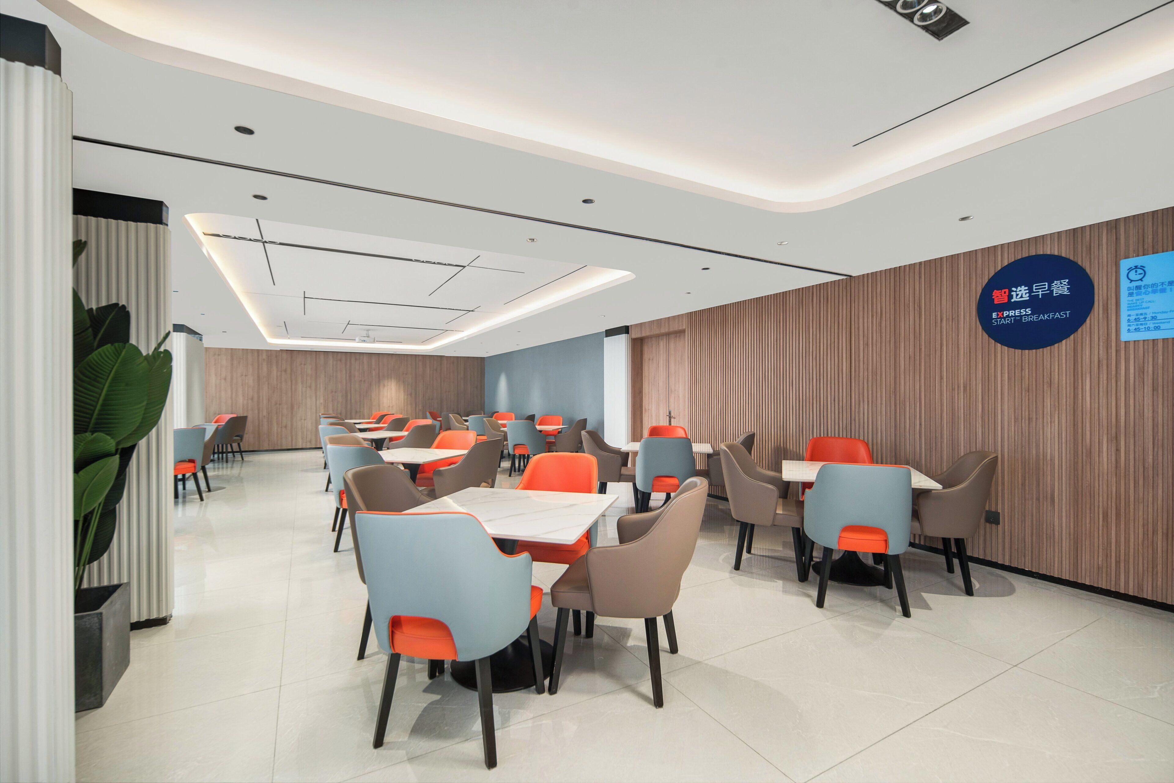A modern dining area with tables and chairs, a sign indicating breakfast service, and a wooden wall.
