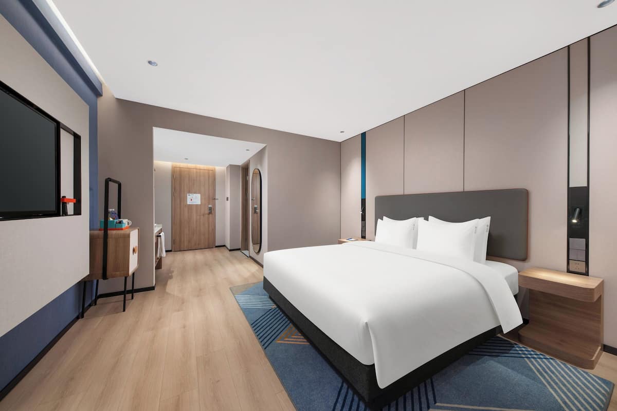 A modern hotel room with a large bed, a bedside table, a mirror, and a view of the hallway.