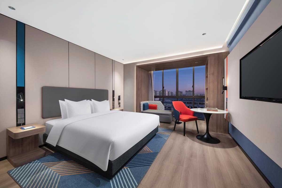 A modern hotel room with a large bed, a desk with a chair, a TV, and a city view.