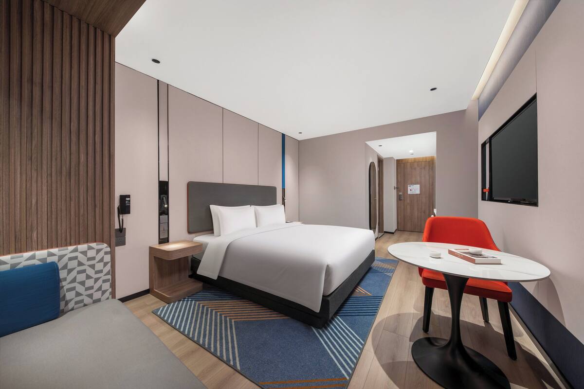 A modern hotel room with a large bed, a desk, a chair, and a TV on the wall.