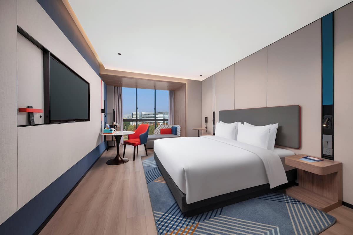 A modern hotel room with a large bed, a desk, a chair, and a view of the city.