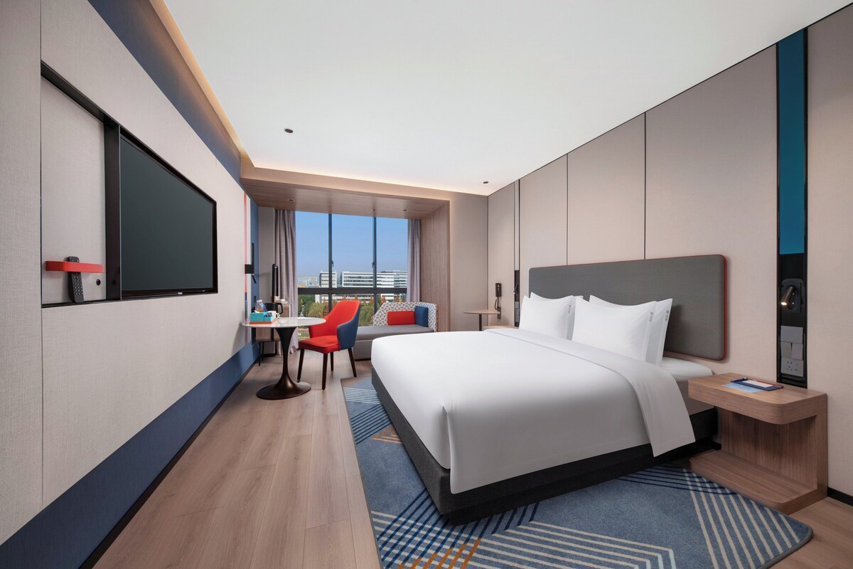 A modern hotel room with a large bed, a desk, a chair, and a view of the city.