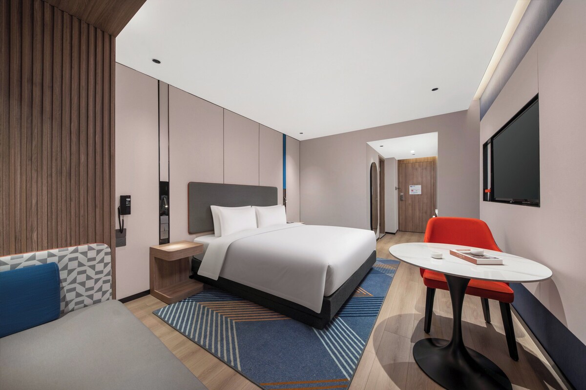 A modern hotel room with a large bed, a desk, a chair, and a TV on the wall.