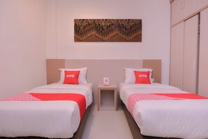 A hotel room with two beds, each with white bedding and red pillows, a small bedside table with a card, and a wall-mounted artwork above the beds. - Oyo 2018 Ring Road Guest House Syariah (Banda Aceh)
