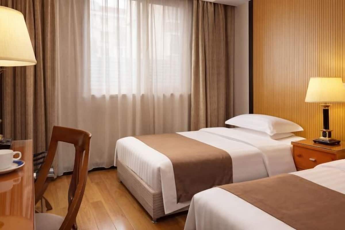A hotel room with two beds, a wooden chair, a table with a cup, and a lamp.