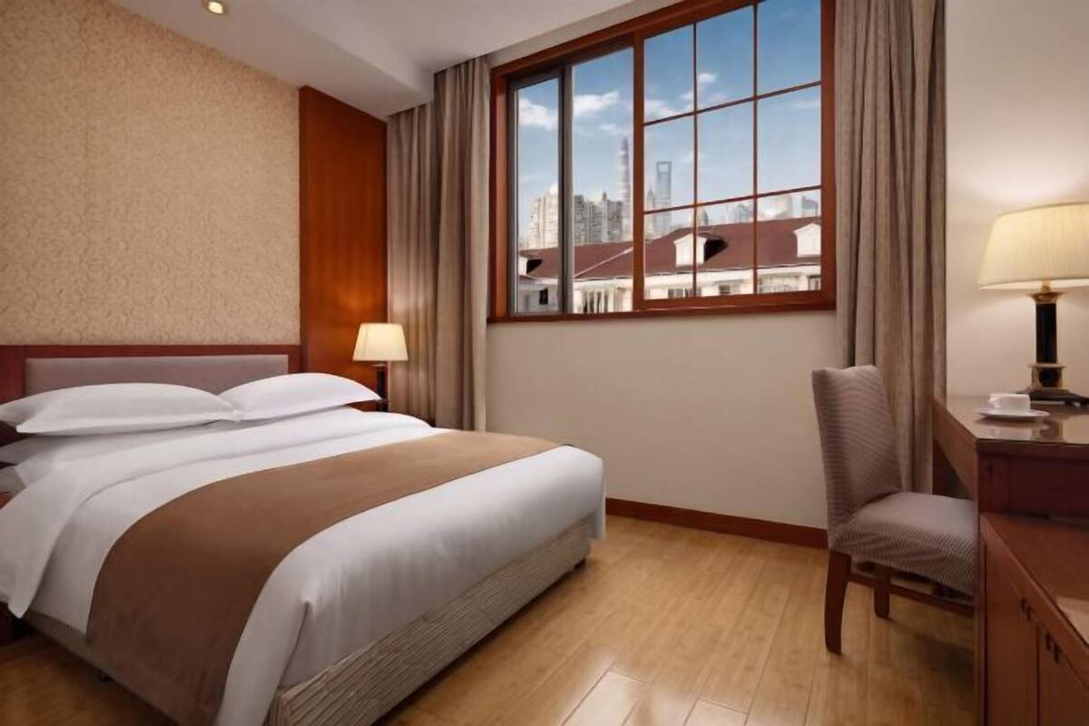 A hotel room with a large bed, a desk with a chair, a window with a city view, and a lamp on the desk.