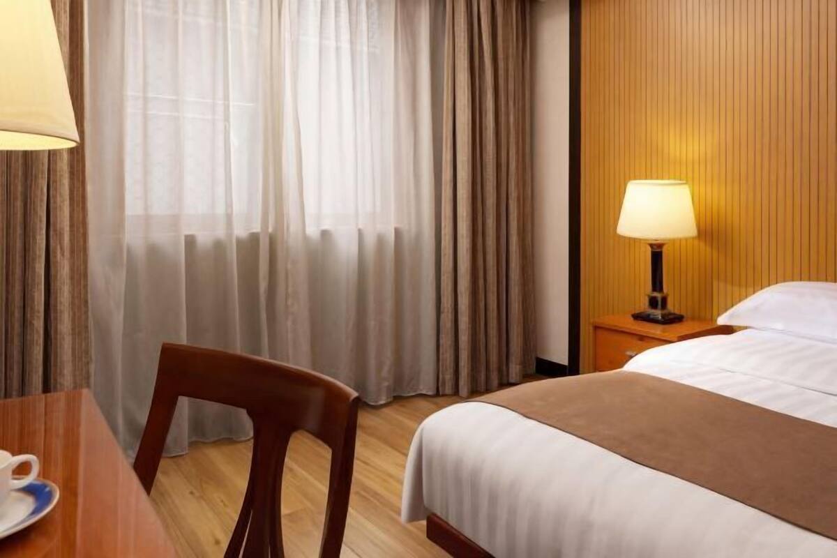 A hotel room with a bed, a chair, a table with a cup, a lamp, and curtains.