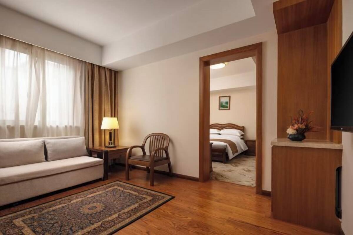 A hotel room with a sofa, chair, bedside table, lamp, and a bed with a wooden headboard.