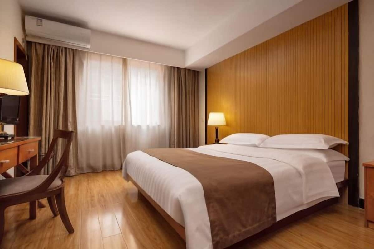 A hotel room with a bed, a desk with a chair, a lamp, and an air conditioning unit.