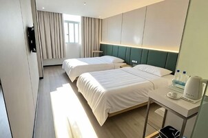 A hotel room with two beds, a bedside table with a kettle, a flat-screen TV, and a window with curtains.