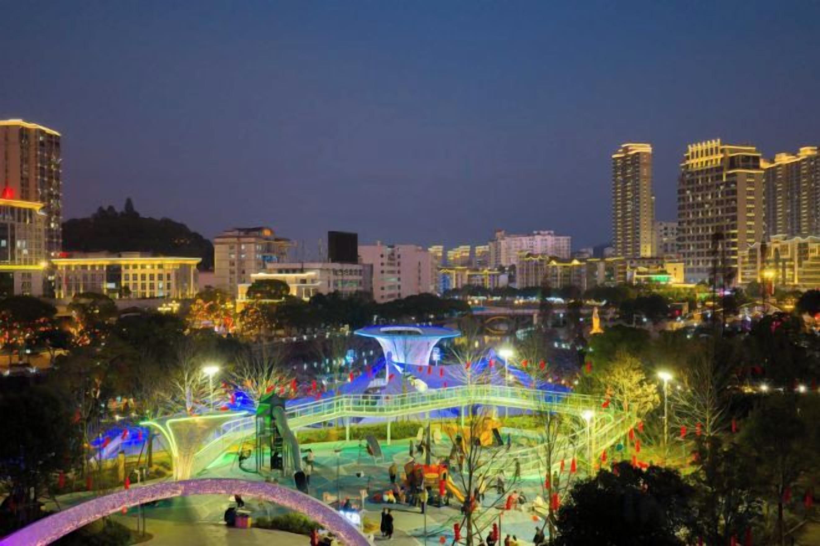 A well-lit urban park with a bridge and pathways, surrounded by high-rise buildings.