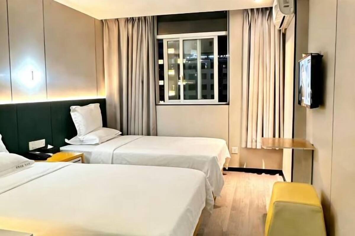 A hotel room with two beds, a bedside table, a lamp, a TV, and a window with curtains.