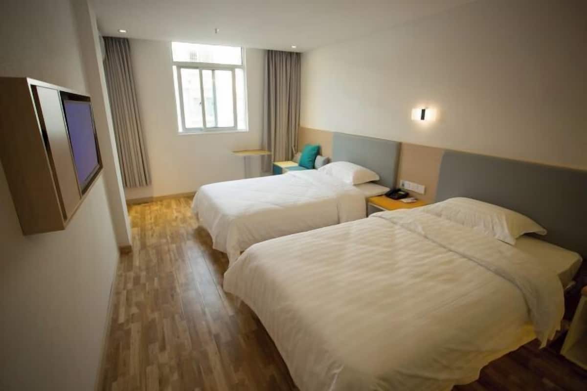 A hotel room with two beds, a window with curtains, a bedside table with a lamp, and a wall-mounted TV.