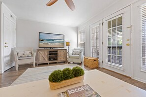 A living room with a television, wicker furniture, a large window with blinds, and a magazine on the floor.