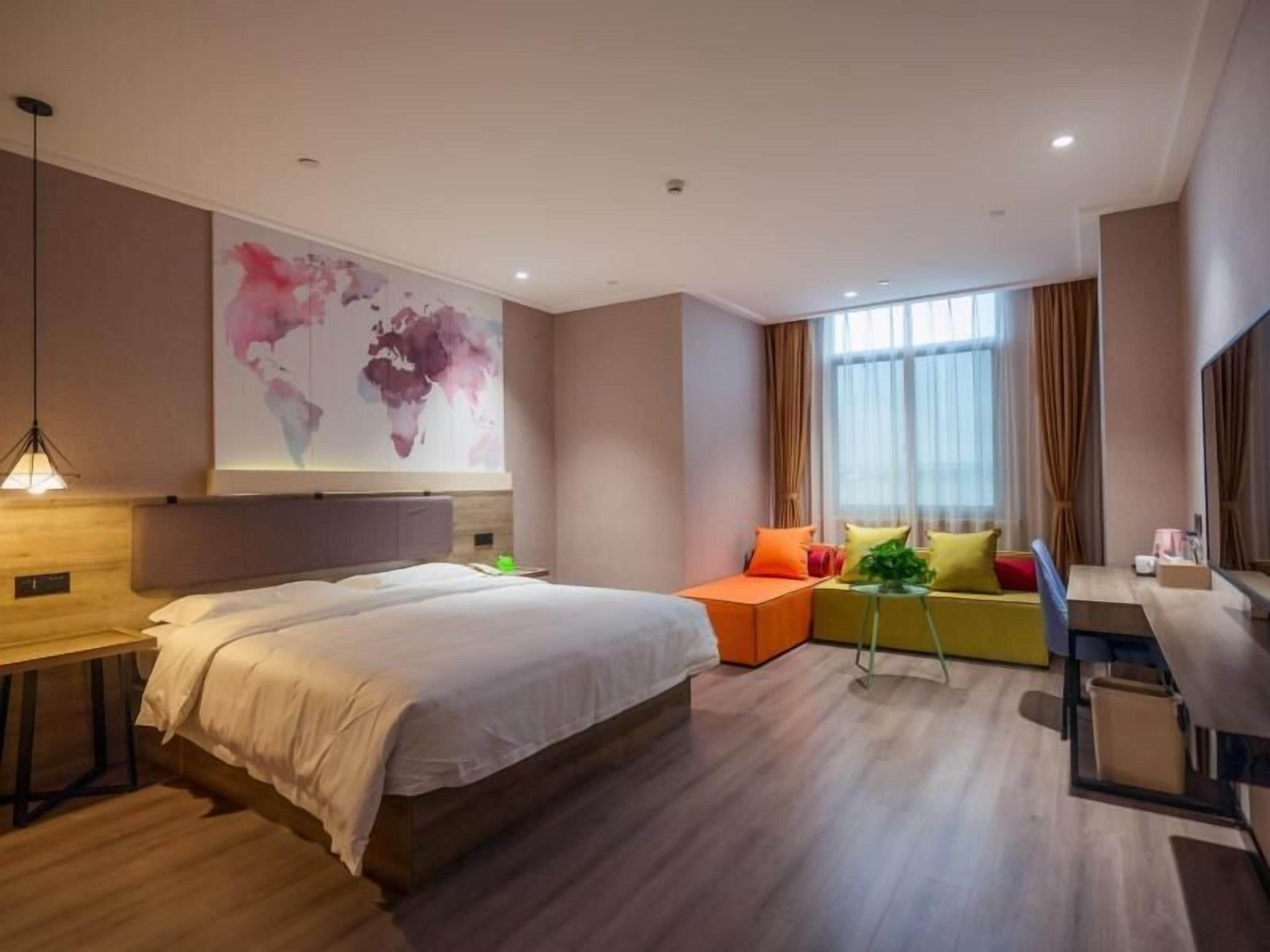 A modern hotel room with a large bed, a seating area with colorful cushions, a desk, and a large abstract wall art piece.