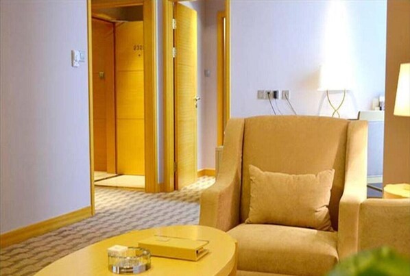 Een hotelkamer met een beige fauteuil, een ronde tafel met een boek en een glas, en een deur die naar een andere kamer leidt.