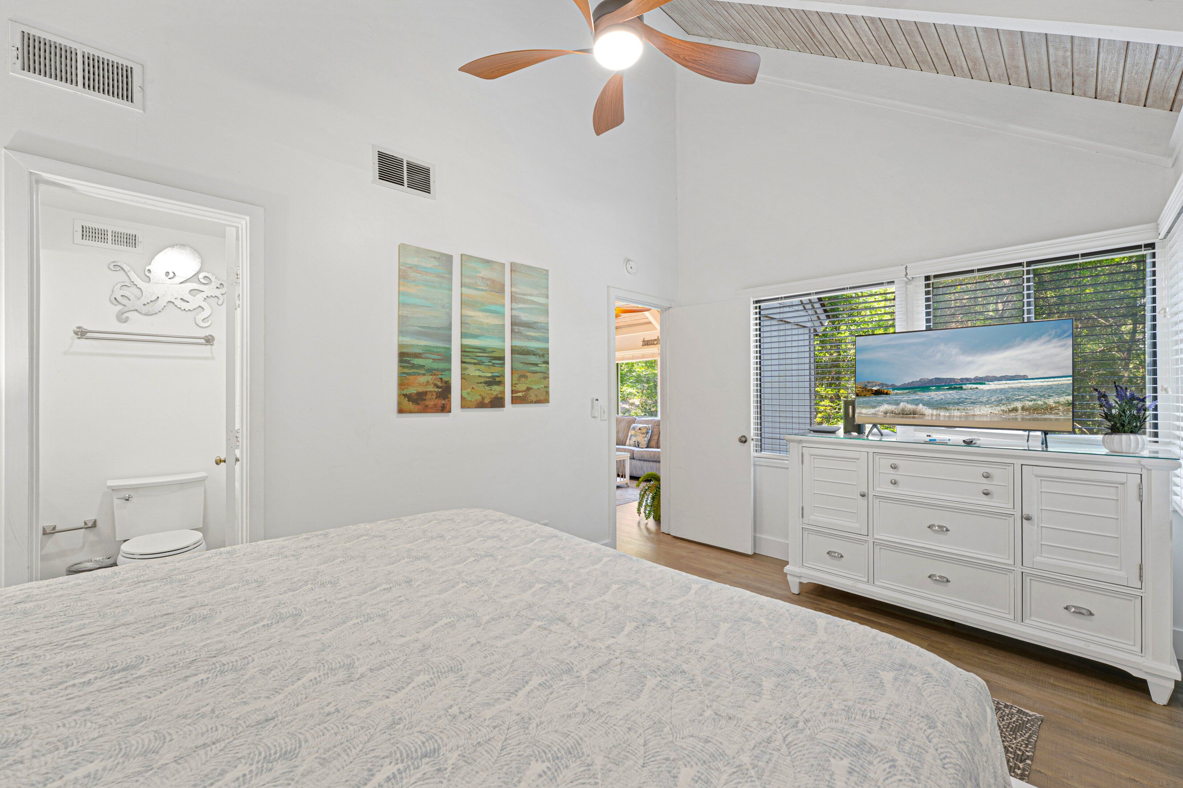 Une chambre à coucher avec un grand lit, un ventilateur de plafond, une commode blanche et un téléviseur.