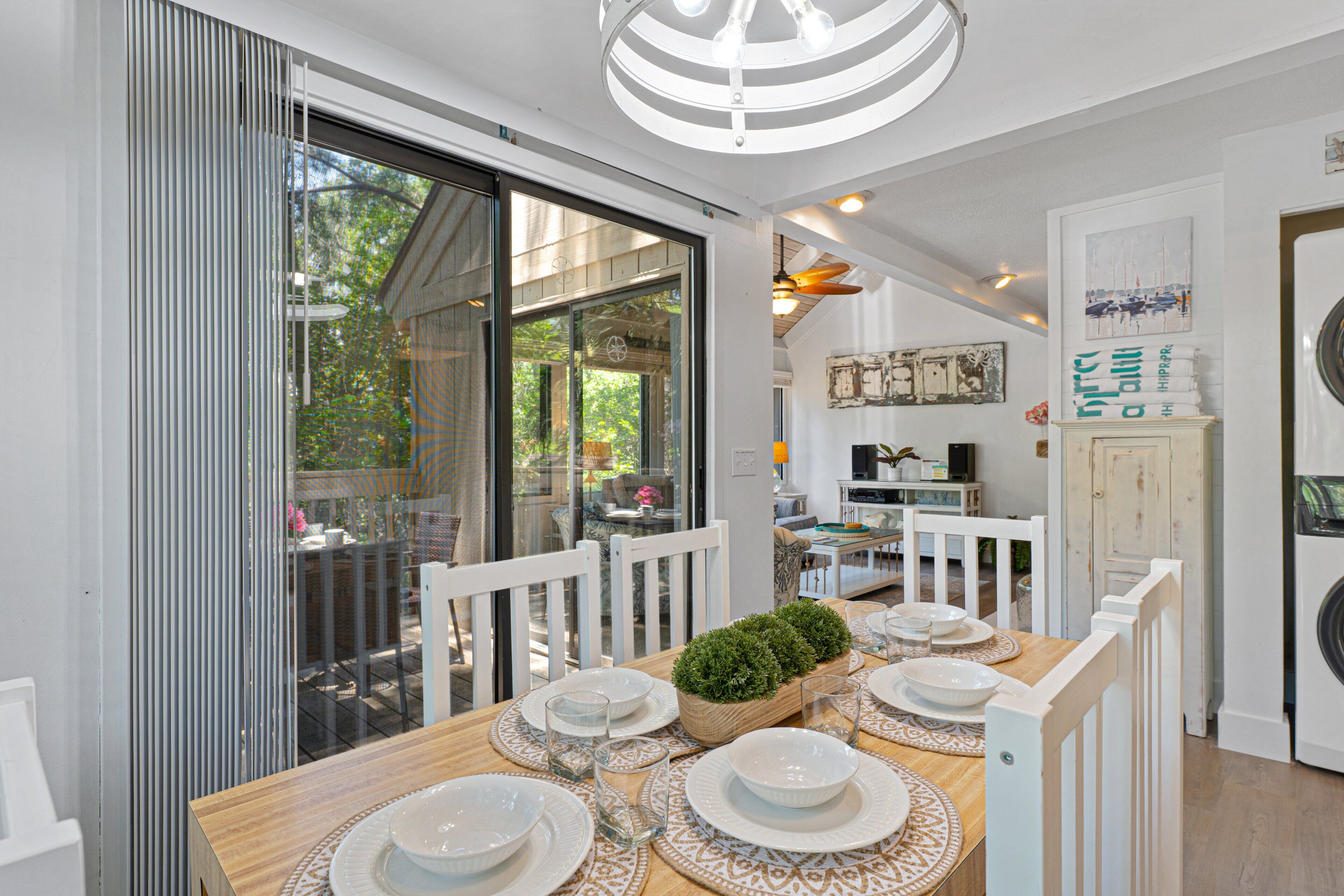 A dining area with a wooden table set for six, surrounded by white chairs, with a view of the outdoors through large sliding glass doors.
