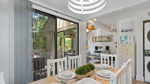 A dining area with a wooden table set for six, surrounded by white chairs, with a view of the outdoors through large sliding glass doors.