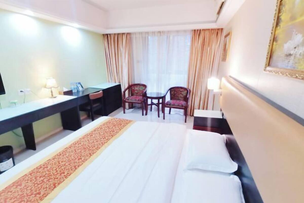 Une chambre d’hôtel avec un lit, un bureau, deux chaises, une petite table et une fenêtre avec des rideaux.