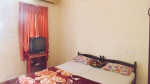 A single bed with a floral bedspread, a television on a stand, and a window with curtains. - Chalet Regency Homestay (Ambalapuzha)