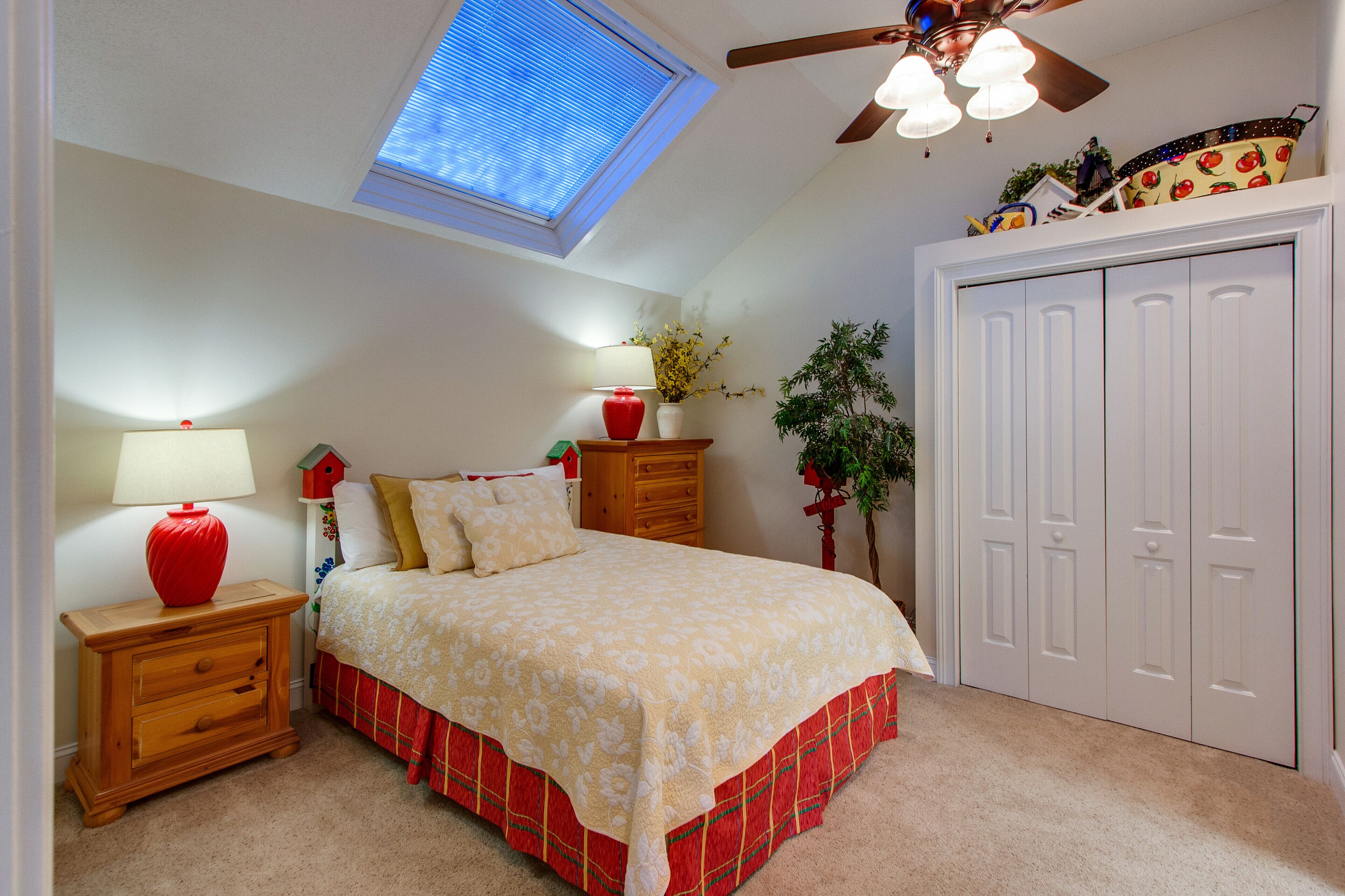 A bedroom with a skylight, a bed with a red skirt, a wooden nightstand with a red lamp, a dresser, and a white wardrobe.