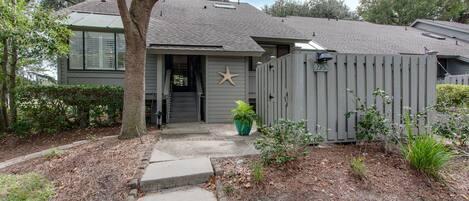 Una casa de dos pisos con fachada gris, un adorno de estrella de mar en la puerta principal y un patio trasero cercado con plantas.