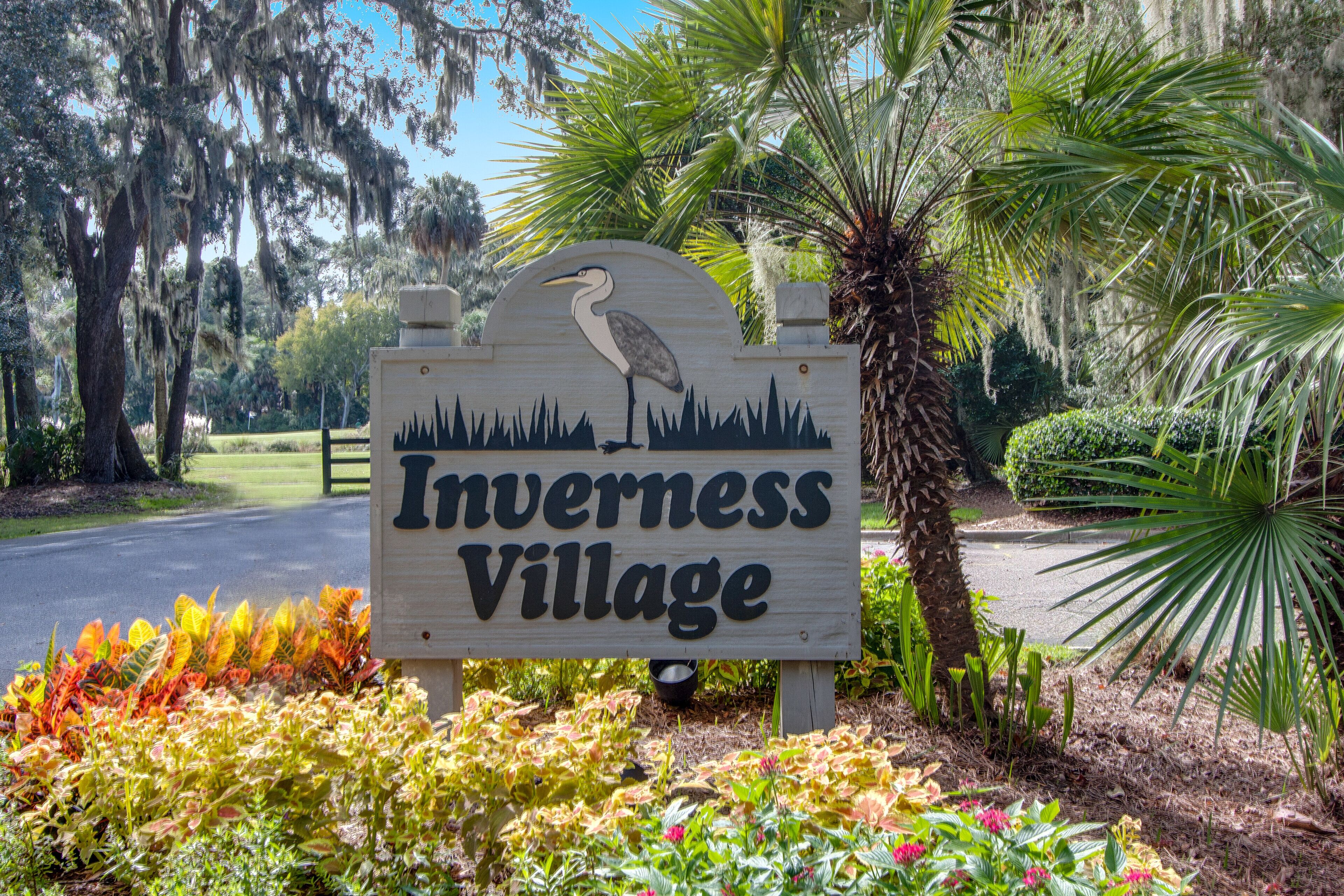 A sign for Inverness Village with a heron graphic, surrounded by lush greenery and a paved road.