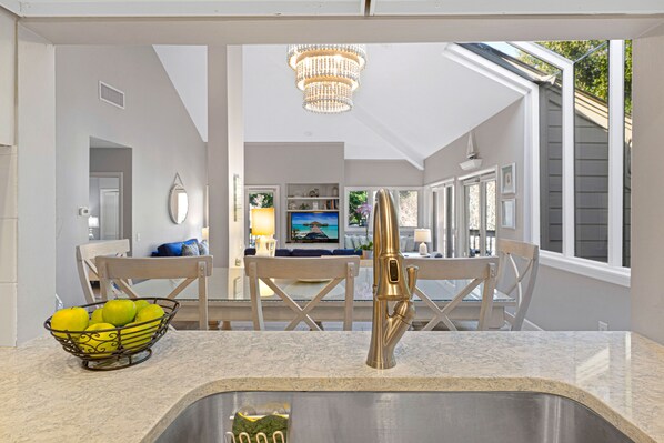 A kitchen with a marble countertop, a stainless steel sink, and a bowl of lemons. There is a chandelier above the sink and a window with a view of the outside.