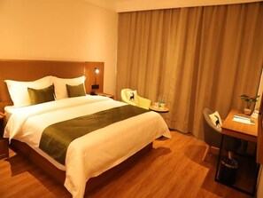 A hotel room with a large bed, a desk, a chair, a small table with a plant, and a curtain.