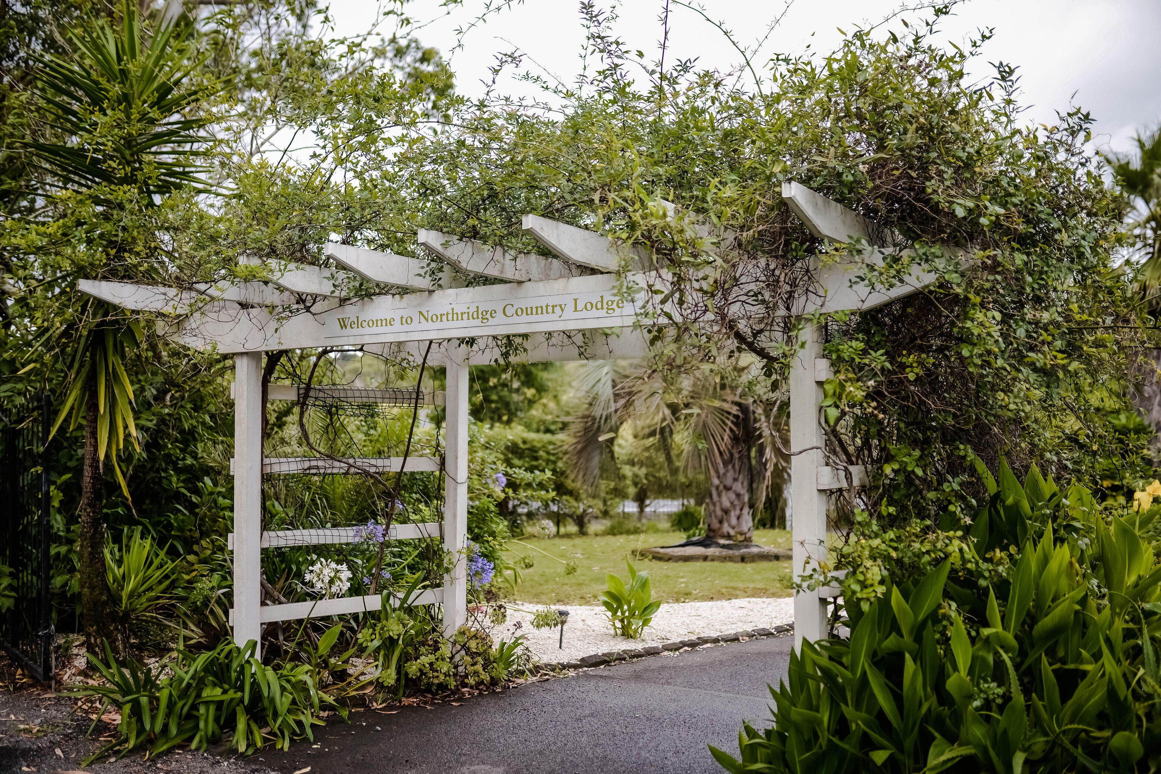 Un arco de madera blanca con el texto "Welcome to Northridge Country Lodge" que da paso a un exuberante jardín.