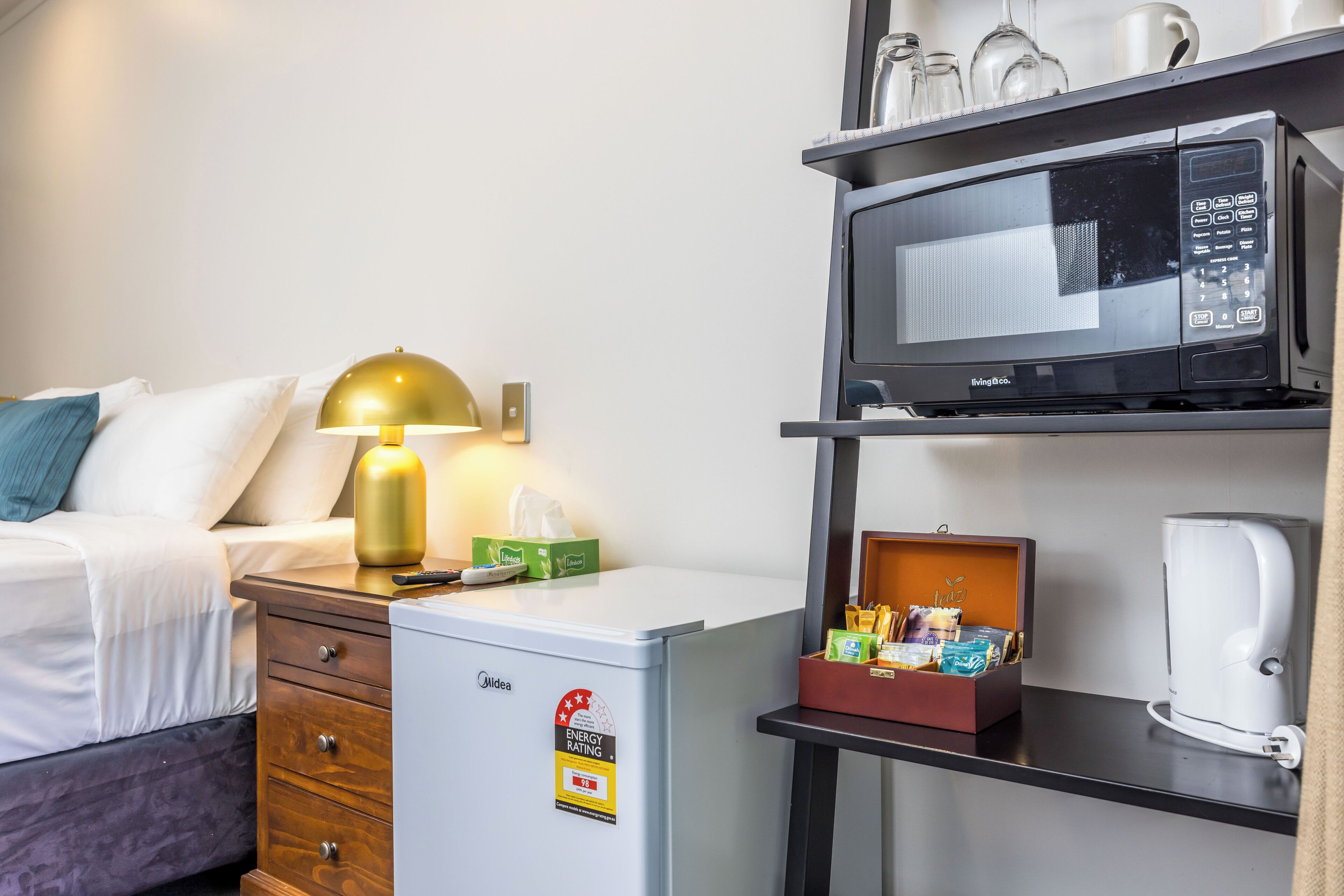 A hotel room with a bed, bedside table with a lamp, a mini-fridge, a microwave, a coffee maker, and a small cabinet with snacks.
