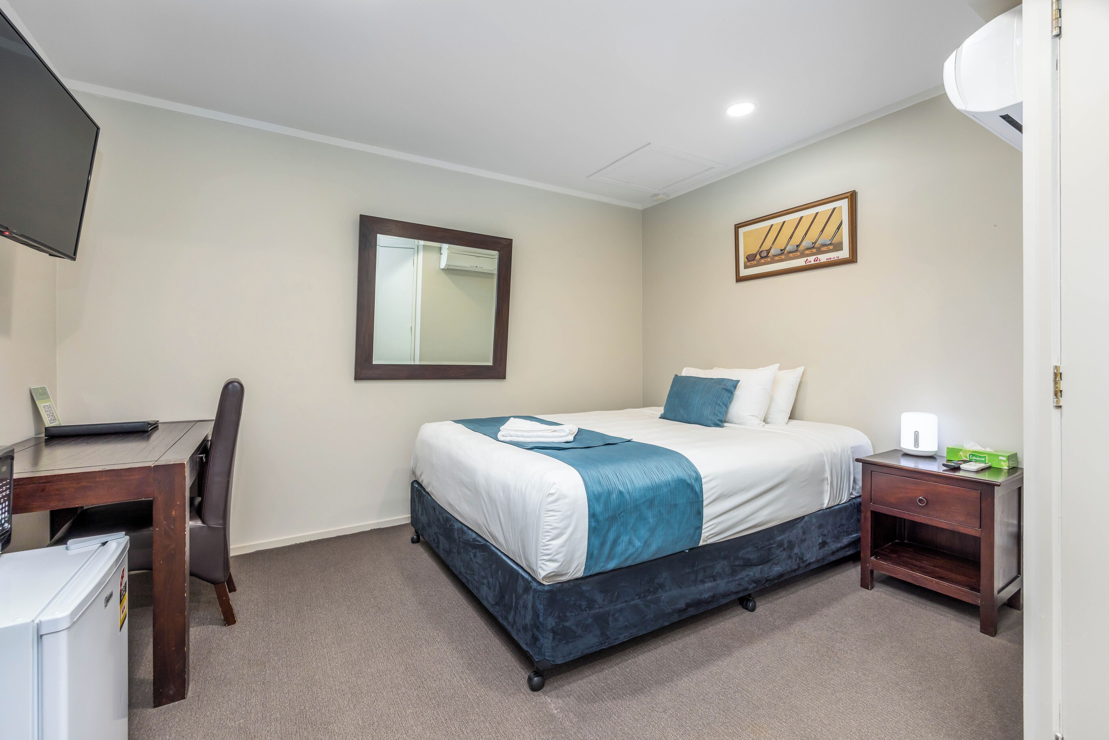 A hotel room with a bed, a desk, a chair, a small table, a nightstand, a mirror, and a wall-mounted TV.