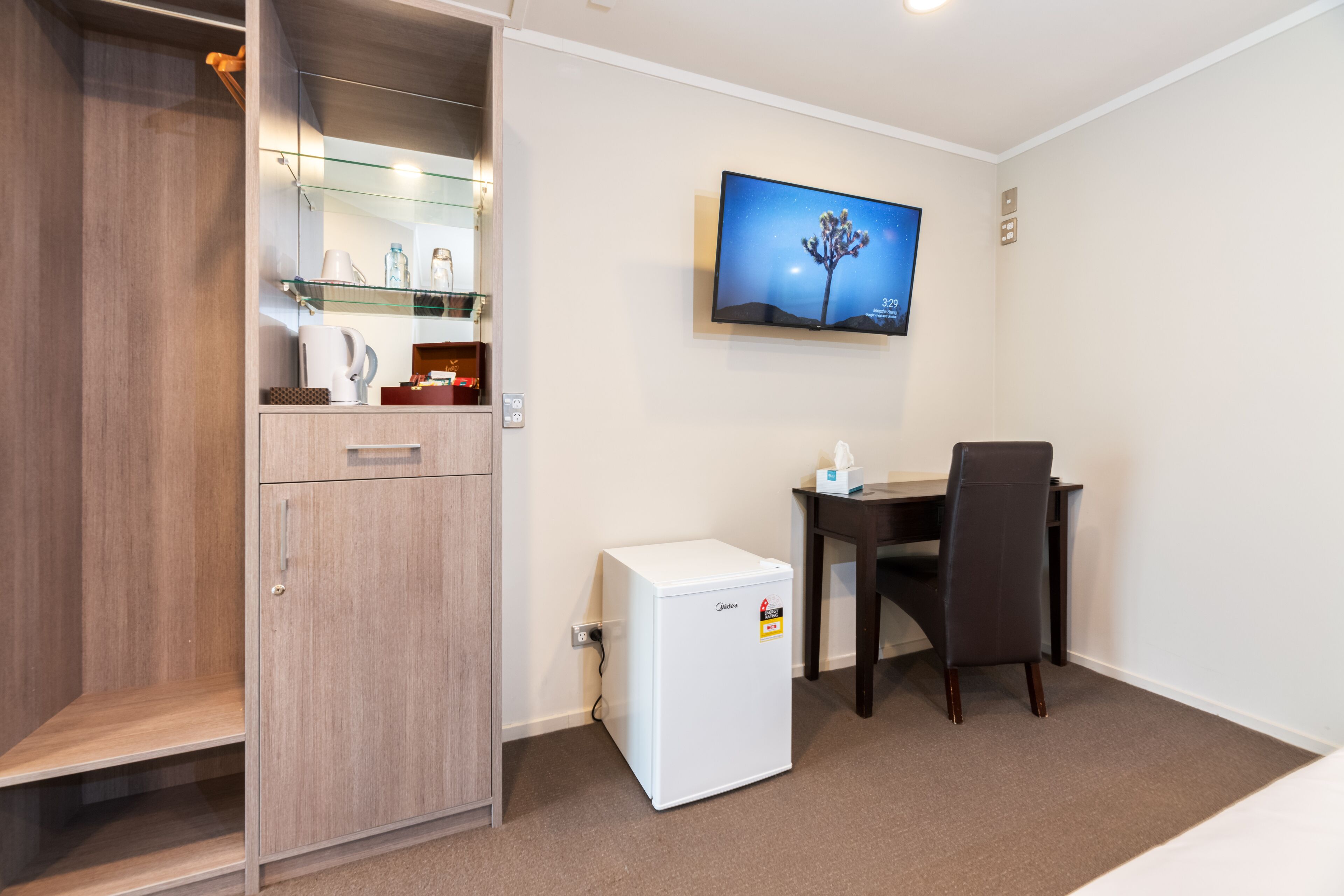 A compact hotel room with a kitchenette, a small refrigerator, a desk with a chair, a flat-screen TV mounted on the wall, and a wooden wardrobe.